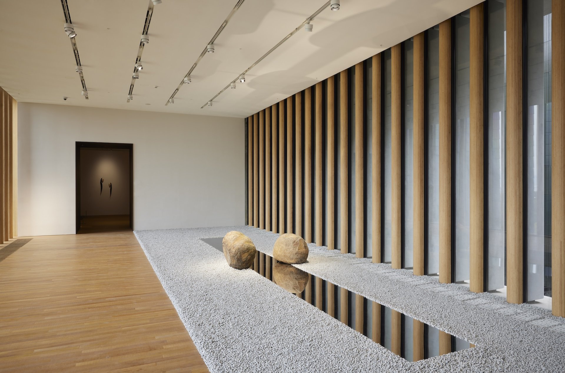 Grey pebbles carpet half of the wooden floor along the length of a corridor gallery. On the pebbles, a long stainless-steel plate with a mirror surface stretches across the room with two stones placed opposite each other at the centre. The steel plate reflects the surrounding environment.